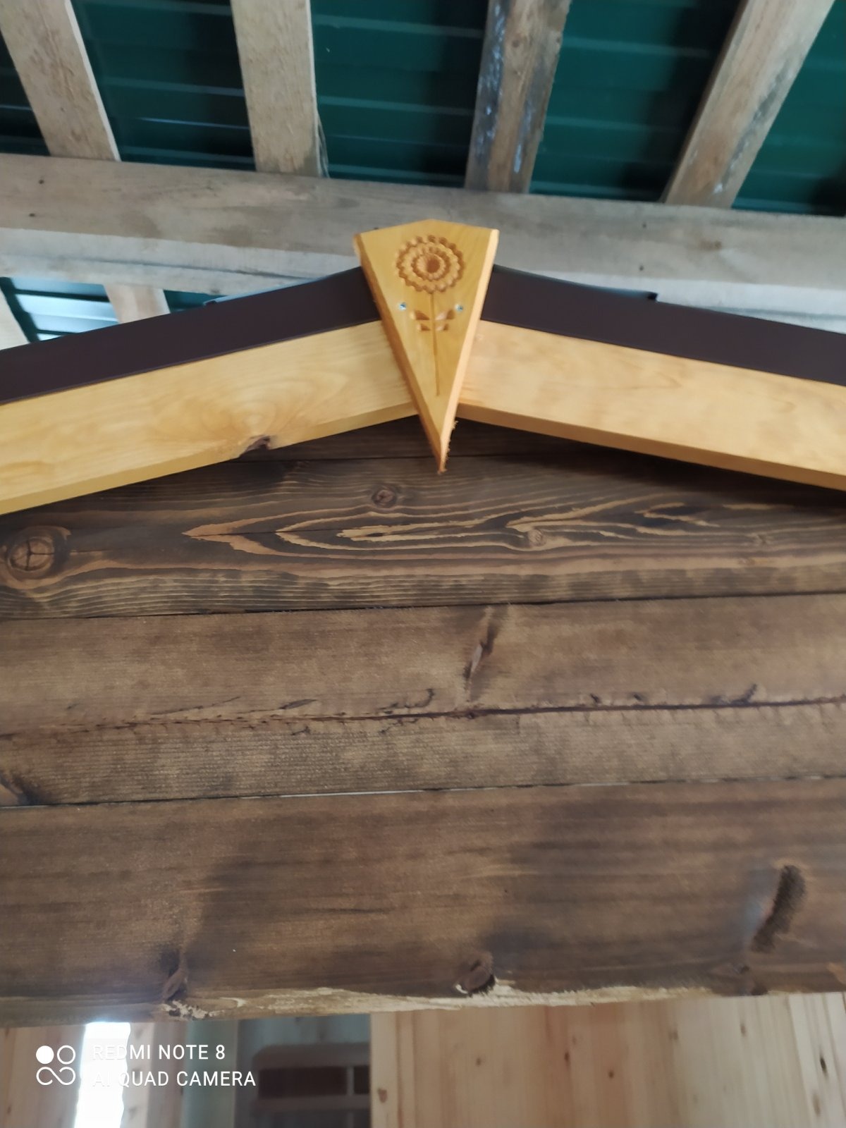 Detal drewnianej konstrukcji z dekoracyjnym elementem w kształcie kwiatu słonecznika, ciemnobrązowe deski i jasne drewno, widoczna część dachu z zieloną blachą.