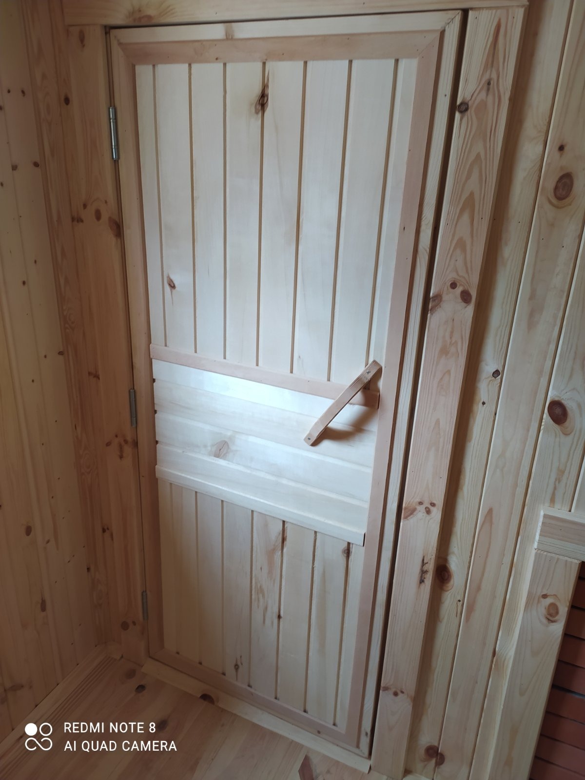 Wnętrze sauny w trakcie budowy: widoczna drewniana konstrukcja ścian, drzwi z ukośną klamką i niedokończone wykończenie.