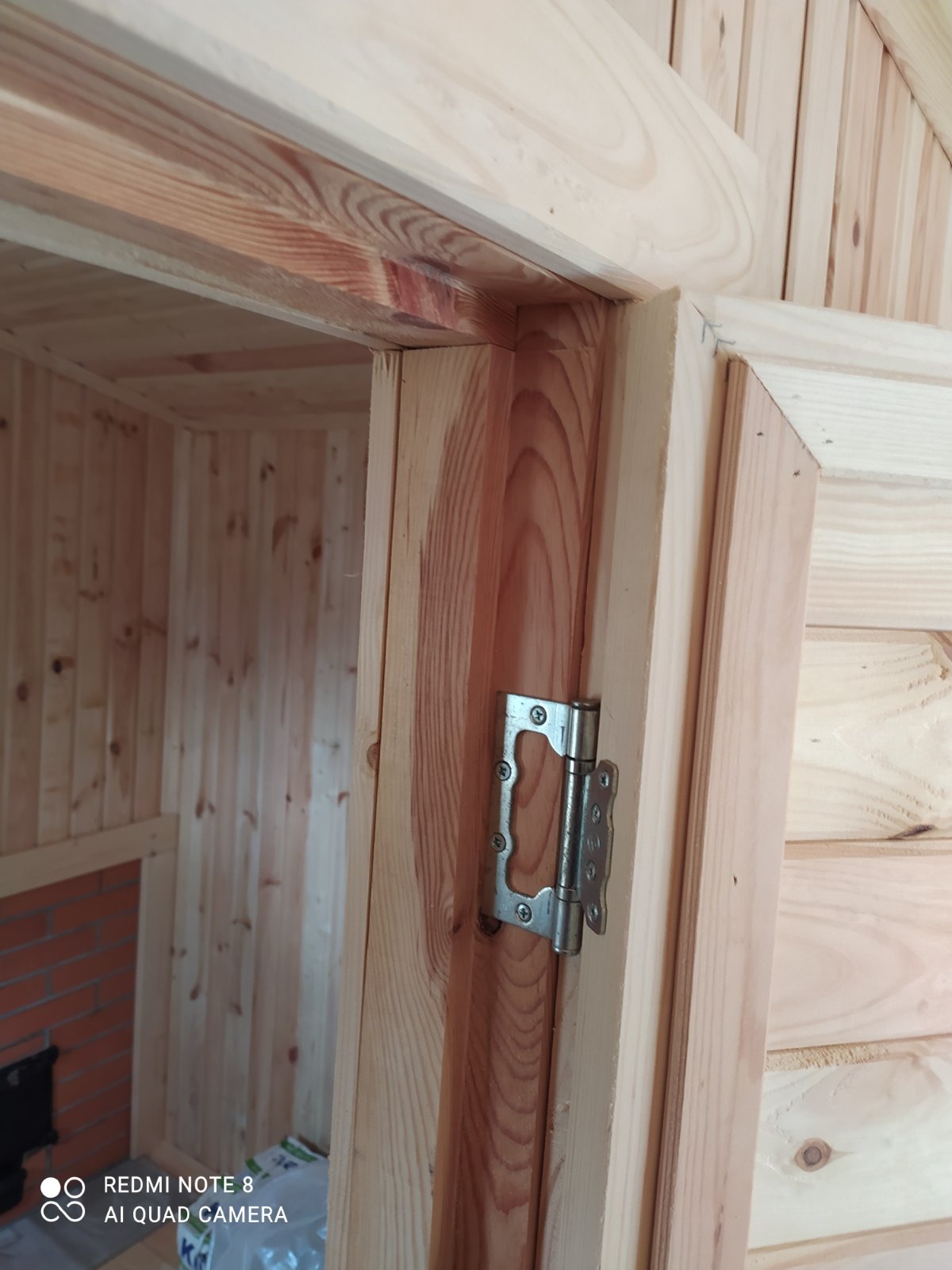 Fragment drewnianej konstrukcji z zawiasem drzwiowym, widoczny mur z cegły w tle.