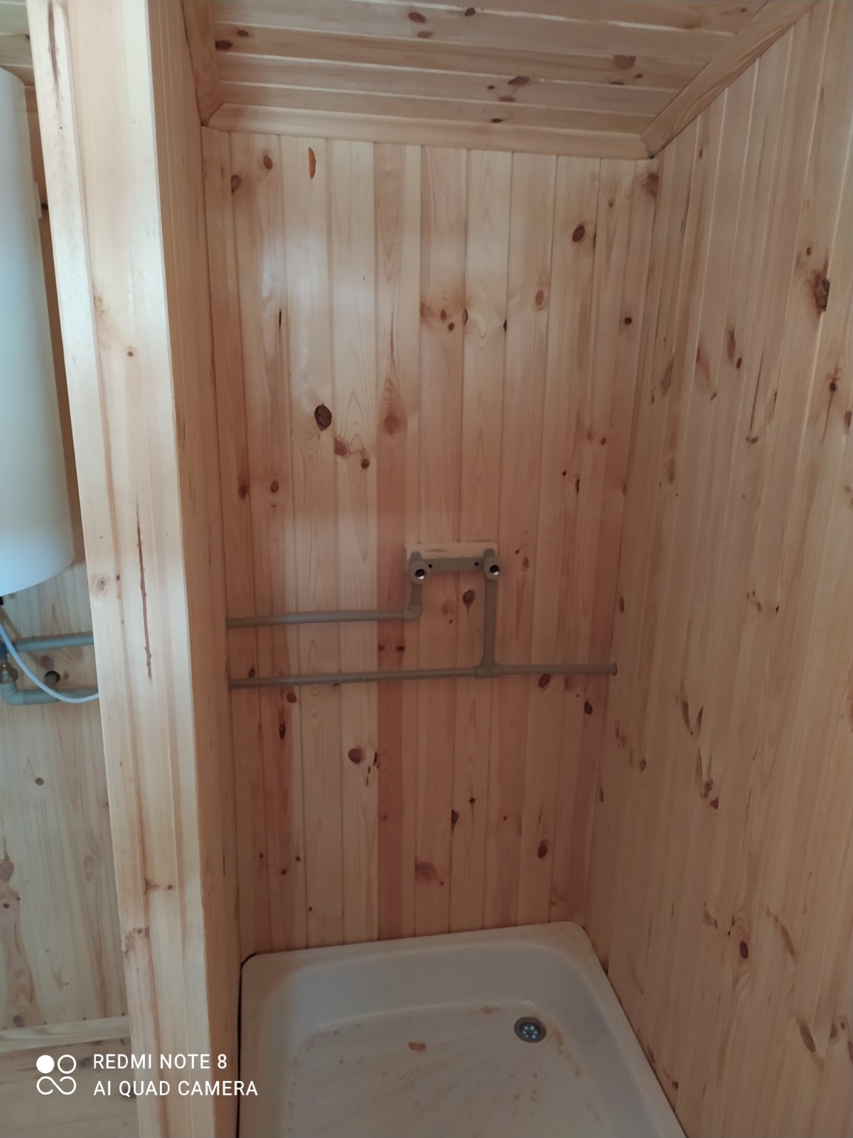 Wnętrze kabiny prysznicowej wykończonej boazerią, z widocznymi rurami instalacji wodnej i białym brodzikiem.