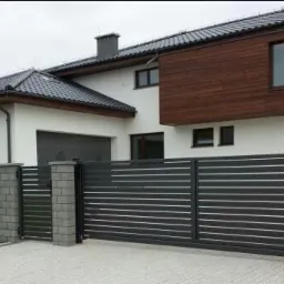 Nowoczesna, pozioma, metalowa brama wjazdowa i furtka w kolorze antracytowym, z elementami murowanymi z szarej kostki, na tle białego domu z drewnianą elewacją i szarą dachówką.