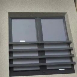 Okna zabezpieczone poziomymi, metalowymi prętami ochronnymi, widok z dołu na elewację budynku o szarej, ziarnistej fakturze.