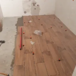 Podłoga w trakcie układania płytek imitujących drewno, z użyciem poziomicy i systemu poziomowania z pomarańczowymi klipsami, widoczne narzędzia i worki z elementami systemu.