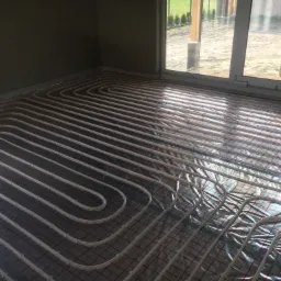 Białe rury ogrzewania podłogowego ułożone w pętle na folii z czerwoną siatką, widoczne przez okno elementy ogrodu.