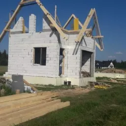 Surowy stan domu jednorodzinnego z białych bloczków, widoczne drewniane elementy konstrukcji dachu, okno bez ramy, na trawie leżą deski, w tle inne domy.