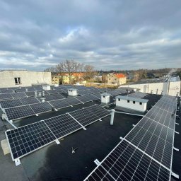 Zuzia Basa - Widok z góry na instalację paneli słonecznych na płaskim dachu budynku, widoczne narzędzia monterskie i fragment buta osoby instalującej, w tle zabudowa miejska pod pochmurnym niebem.