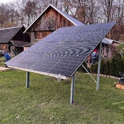 Zuzia Basa - Montaż paneli fotowoltaicznych na metalowej konstrukcji na trawniku, w tle drewniane budynki gospodarcze i drzewa bez liści, widoczni dwaj monterzy w trakcie pracy.