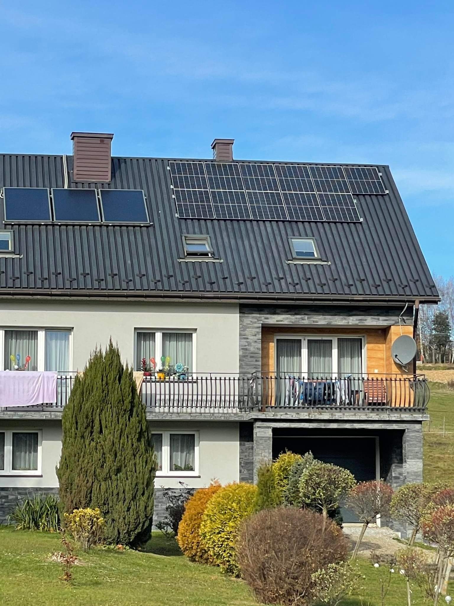 Dwukondygnacyjny dom z ciemnoszarym dachem, na którym zamontowano panele słoneczne i kolektory słoneczne. Na balkonie widoczne dekoracje i suszące się pranie. Ogród z różnorodną roślinnością.