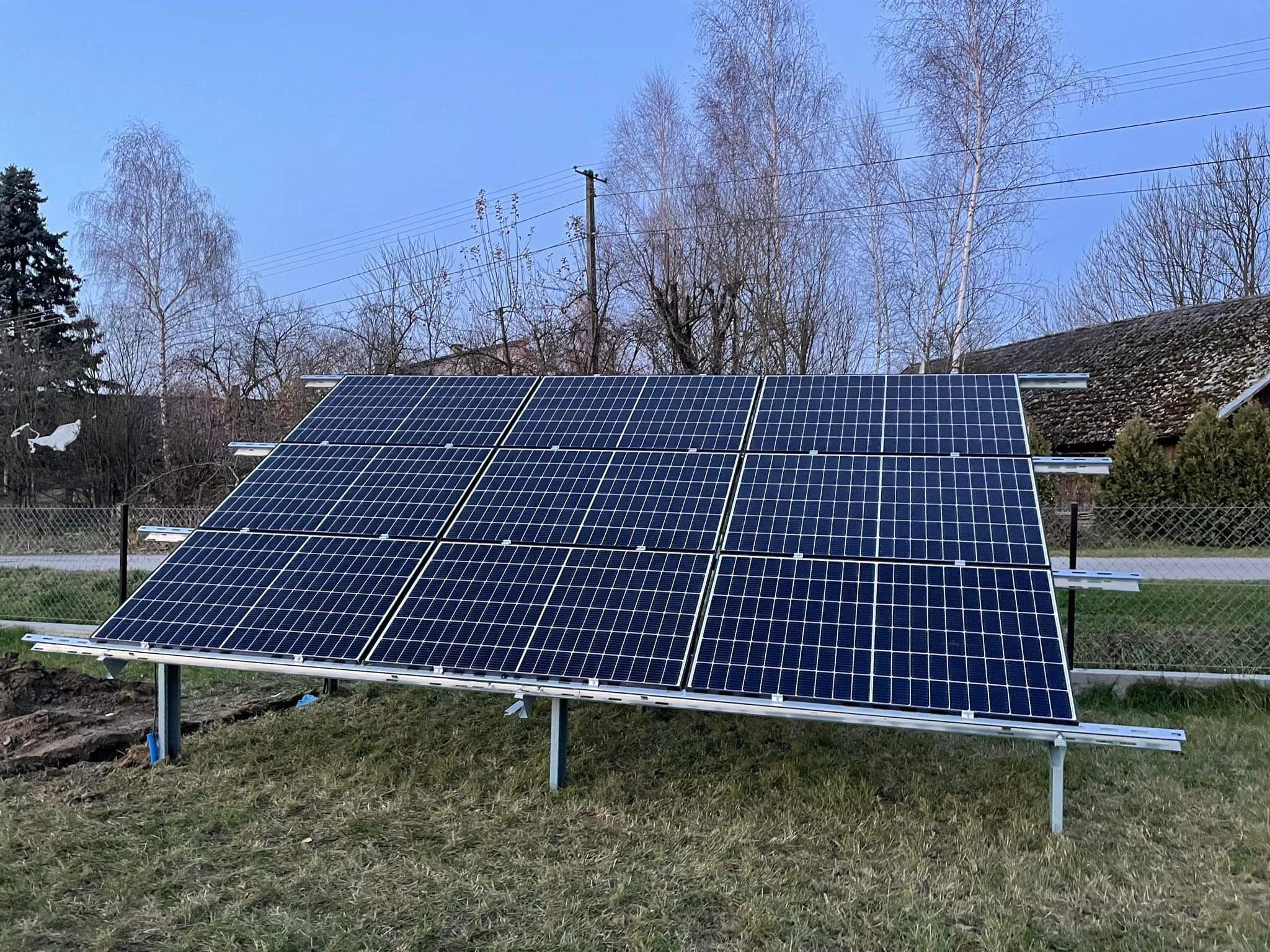 Rząd paneli słonecznych na metalowej konstrukcji, widoczne przygotowane wykopy pod kolejne panele, w tle drzewa bez liści i dom z ciemnym dachem.
