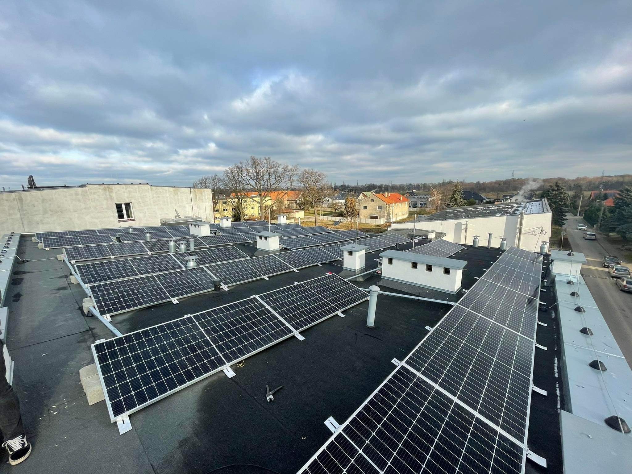 Widok z góry na instalację paneli słonecznych na płaskim dachu budynku, widoczne narzędzia monterskie i fragment buta osoby instalującej, w tle zabudowa miejska pod pochmurnym niebem.