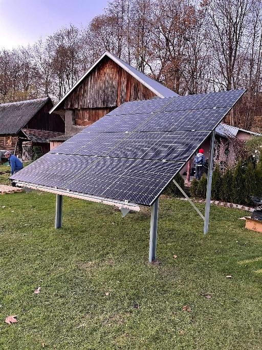 Montaż paneli fotowoltaicznych na metalowej konstrukcji na trawniku, w tle drewniane budynki gospodarcze i drzewa bez liści, widoczni dwaj monterzy w trakcie pracy.
