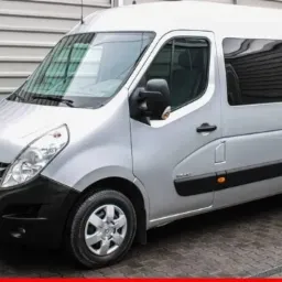 Srebrny bus Renault Master z ciemnymi szybami, zaparkowany przed szarą, metalową ścianą.