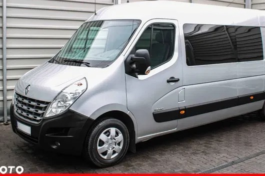 Srebrny bus Renault Master z ciemnymi szybami, zaparkowany przed szarą, metalową ścianą.