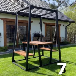 Metalowa konstrukcja huśtawki-ławki z drewnianym blatem i siedziskami na łańcuchach, ustawiona na trawniku przed domem z ciemną dach&oacute;wką.