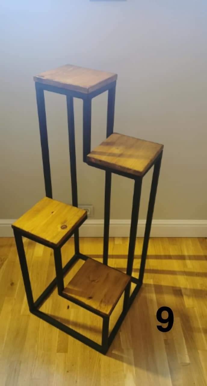 Trzy metalowe kwietniki o różnej wysokości z drewnianymi blatami, ustawione schodkowo na drewnianej podłodze.