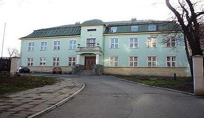 Duży, dwupiętrowy budynek w pastelowym zielonym kolorze z ciemnozielonym dachem, balkonem i schodami wejściowymi, widoczny z perspektywy ulicy z dwoma zaparkowanymi samochodami.