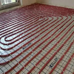 Czerwone rury ogrzewania podłogowego marki Keller ułożone na macie izolacyjnej z siatką, widok z góry, surowy stan wykończenia wnętrza.