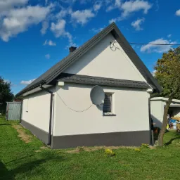 Jednopiętrowy dom z białą elewacją i ciemnoszarą podmurówką, ciemnym dachem z blachodachówki oraz satelitarną anteną na ścianie, na tle błękitnego nieba z chmurami.