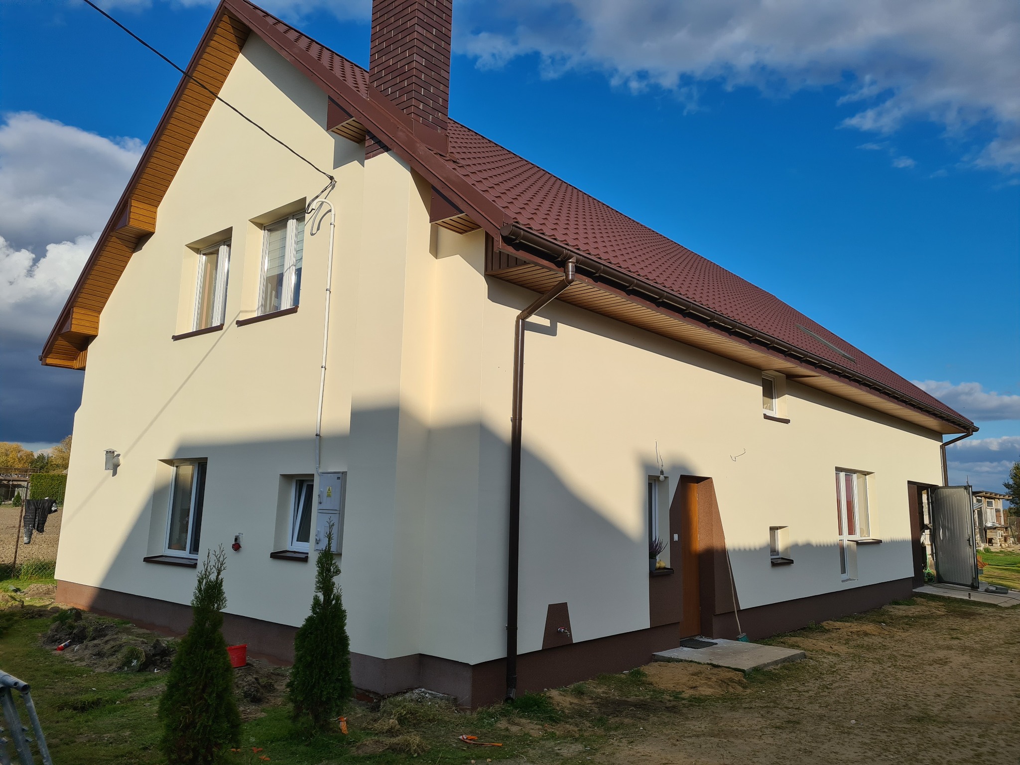 Dom jednorodzinny z beżową elewacją, brązowym dachem i stolarką okienną, widok z zewnątrz na tle błękitnego nieba z chmurami, teren wokół domu częściowo zagospodarowany z małymi drzewkami iglastymi.