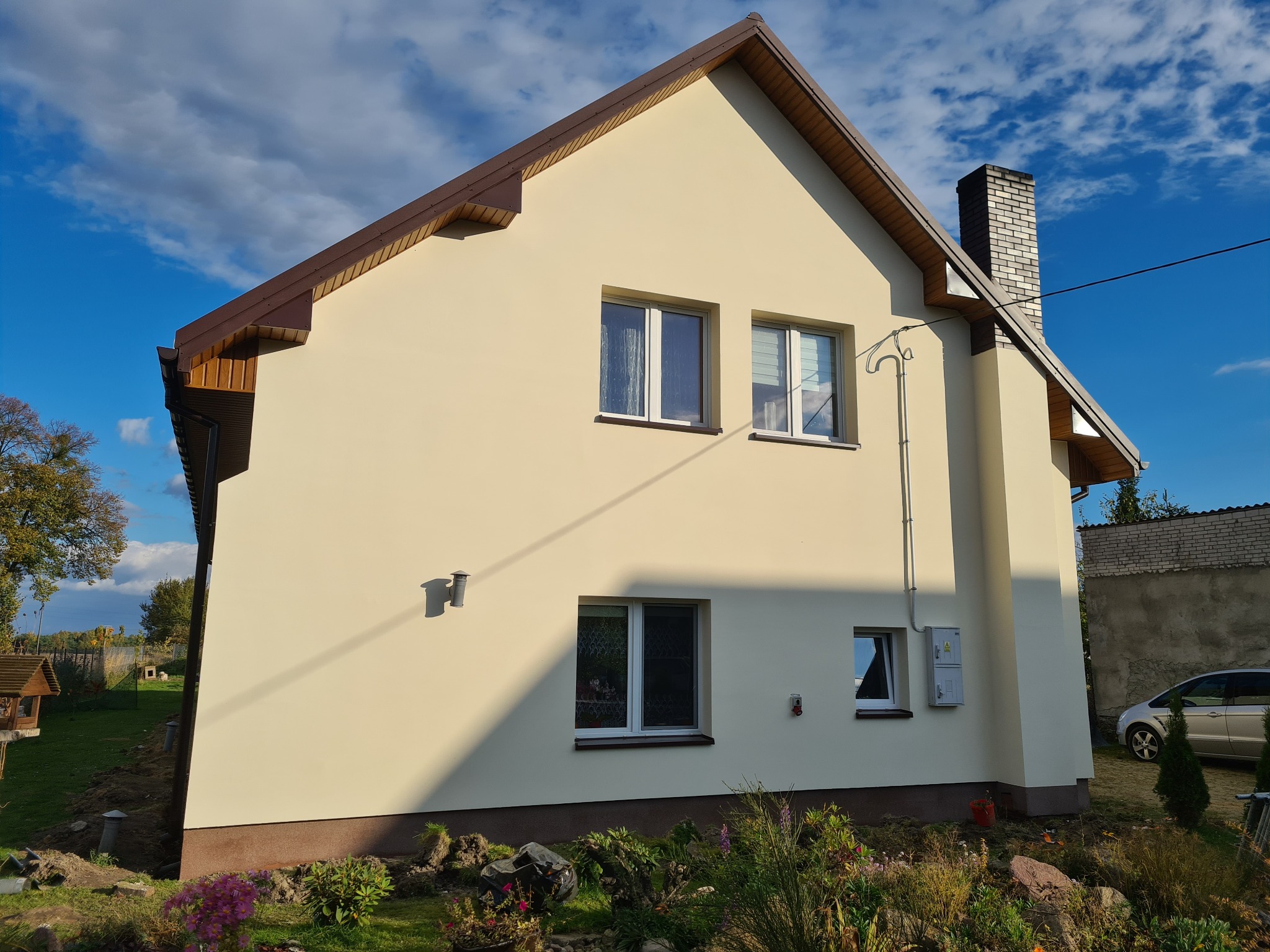 Świeżo otynkowany dom jednorodzinny z brązowym dachem, widok z boku, z widocznymi oknami, kominem z cegły i elementami instalacji na elewacji, ogród z kwiatami na pierwszym planie.