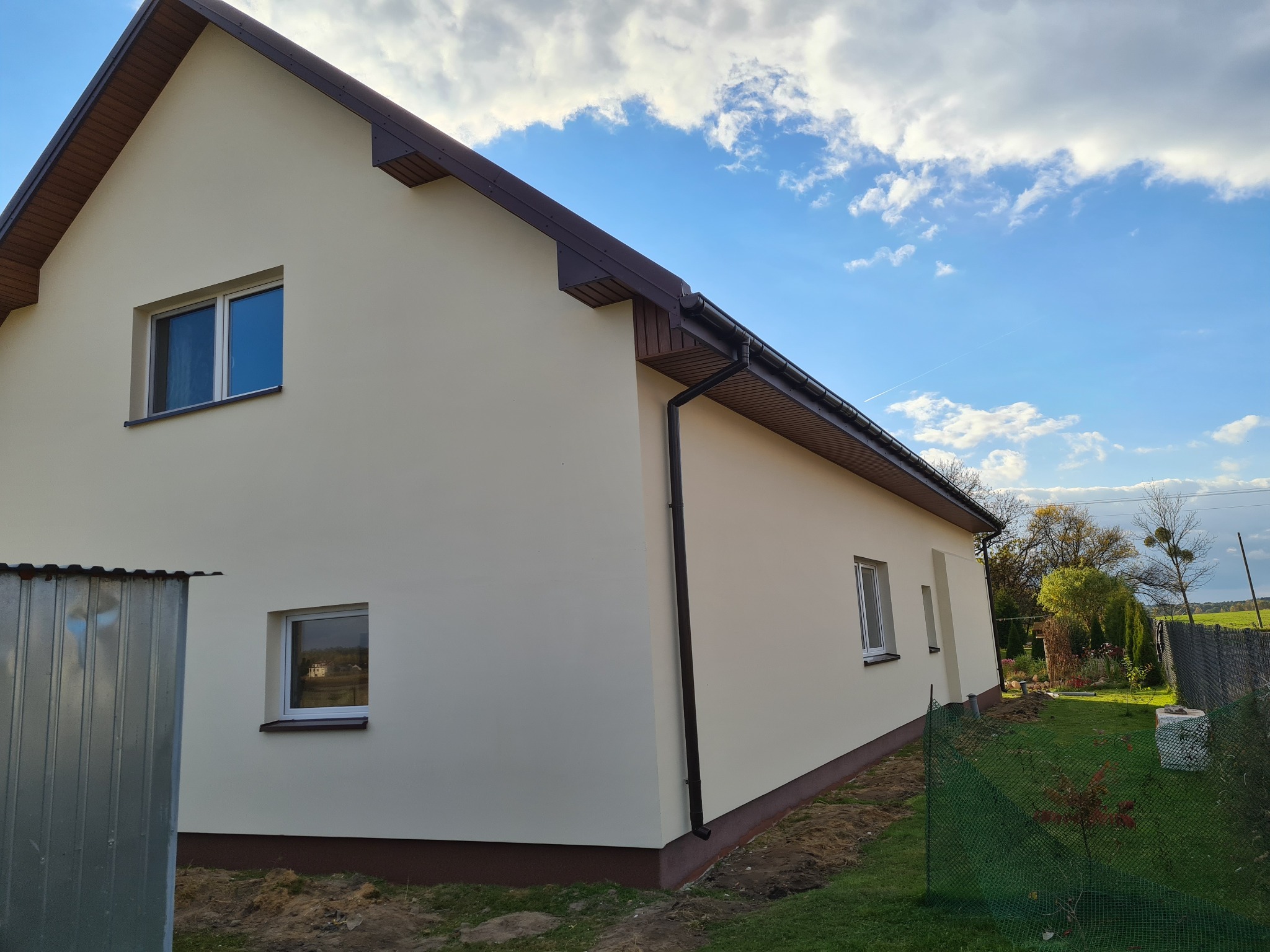 Świeżo pomalowana elewacja domu jednorodzinnego w kolorze kremowym, z brązowymi elementami wykończeniowymi dachu i fundamentów, na tle błękitnego nieba z obłokami.
