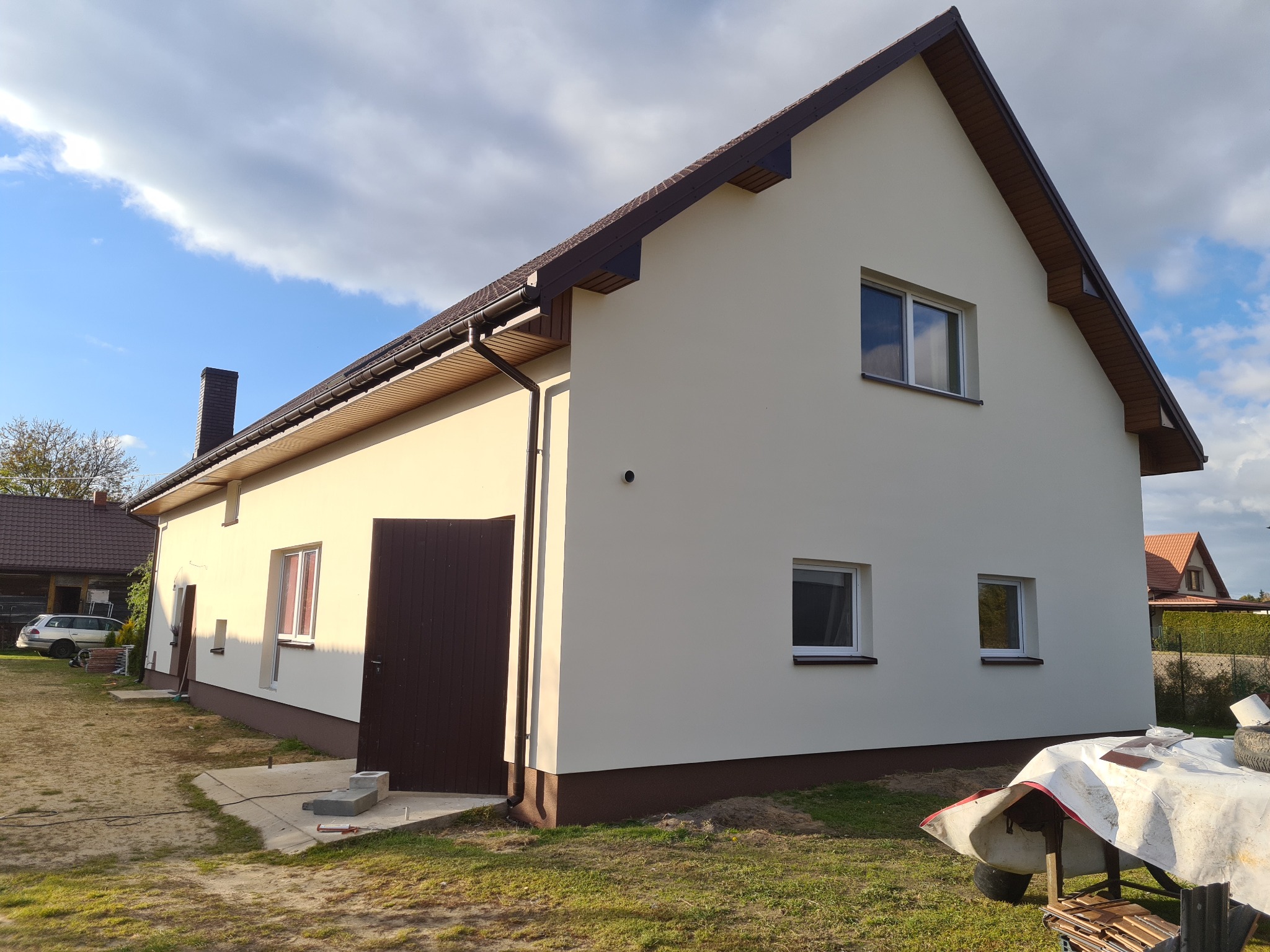 Dwukondygnacyjny dom z jasną elewacją, brązowym dachem i otwartymi drzwiami garażowymi, widoczny w słoneczny dzień z lekkim zachmurzeniem.