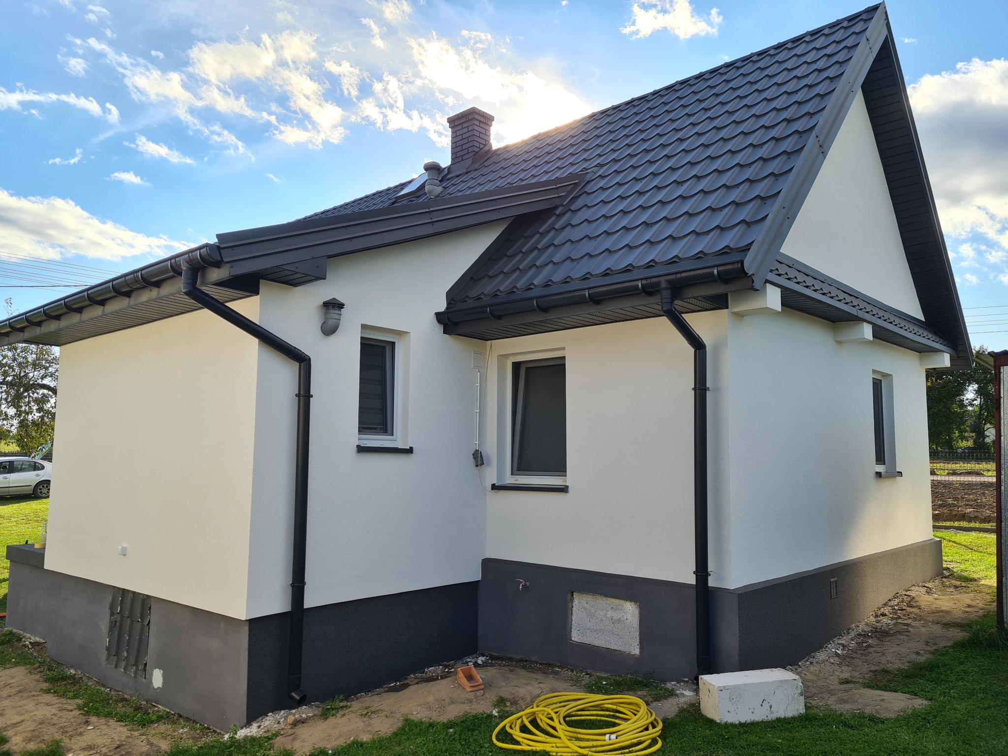 Świeżo pomalowany dom jednorodzinny z ciemną podmurówką, grafitową blachodachówką i rynnami, żółty wąż ogrodowy leży na trawie.