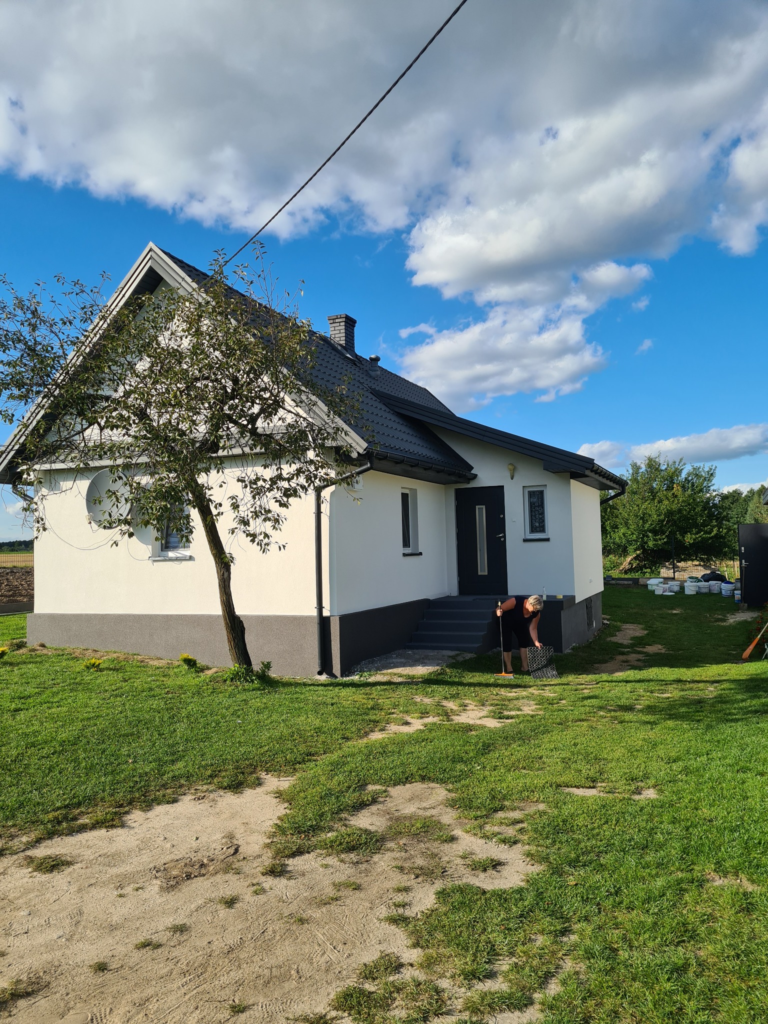 Dom jednorodzinny z białą elewacją i szarym cokołem, na trawniku kobieta zamiatająca liście, błękitne niebo z chmurami w tle.