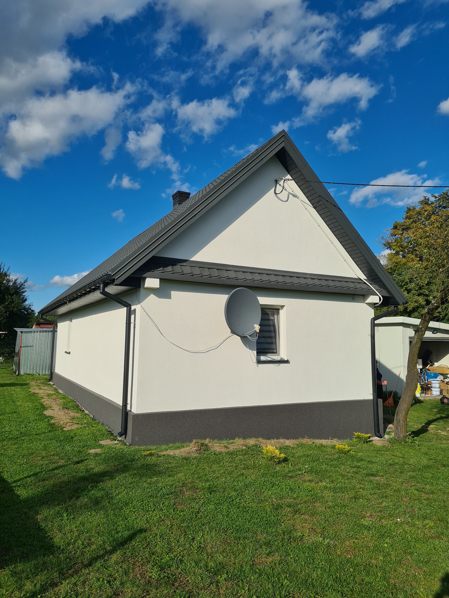 Jednopiętrowy dom z białą elewacją i ciemnoszarą podmurówką, ciemnym dachem z blachodachówki oraz satelitarną anteną na ścianie, na tle błękitnego nieba z chmurami.