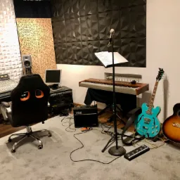 Profesjonalnie wyposażone pomieszczenie: mikser dźwięku, monitory studyjne, pianino, gitary elektryczne i akustyczne, mikrofon na statywie, panele akustyczne na ścianach.