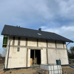 Surowy stan domu jednorodzinnego w trakcie budowy: widoczna czarna dachówka na skośnym dachu z oknem dachowym, zamontowane rynny, ściany z betonu komórkowego i niezabudowany otwór drzwiowy.