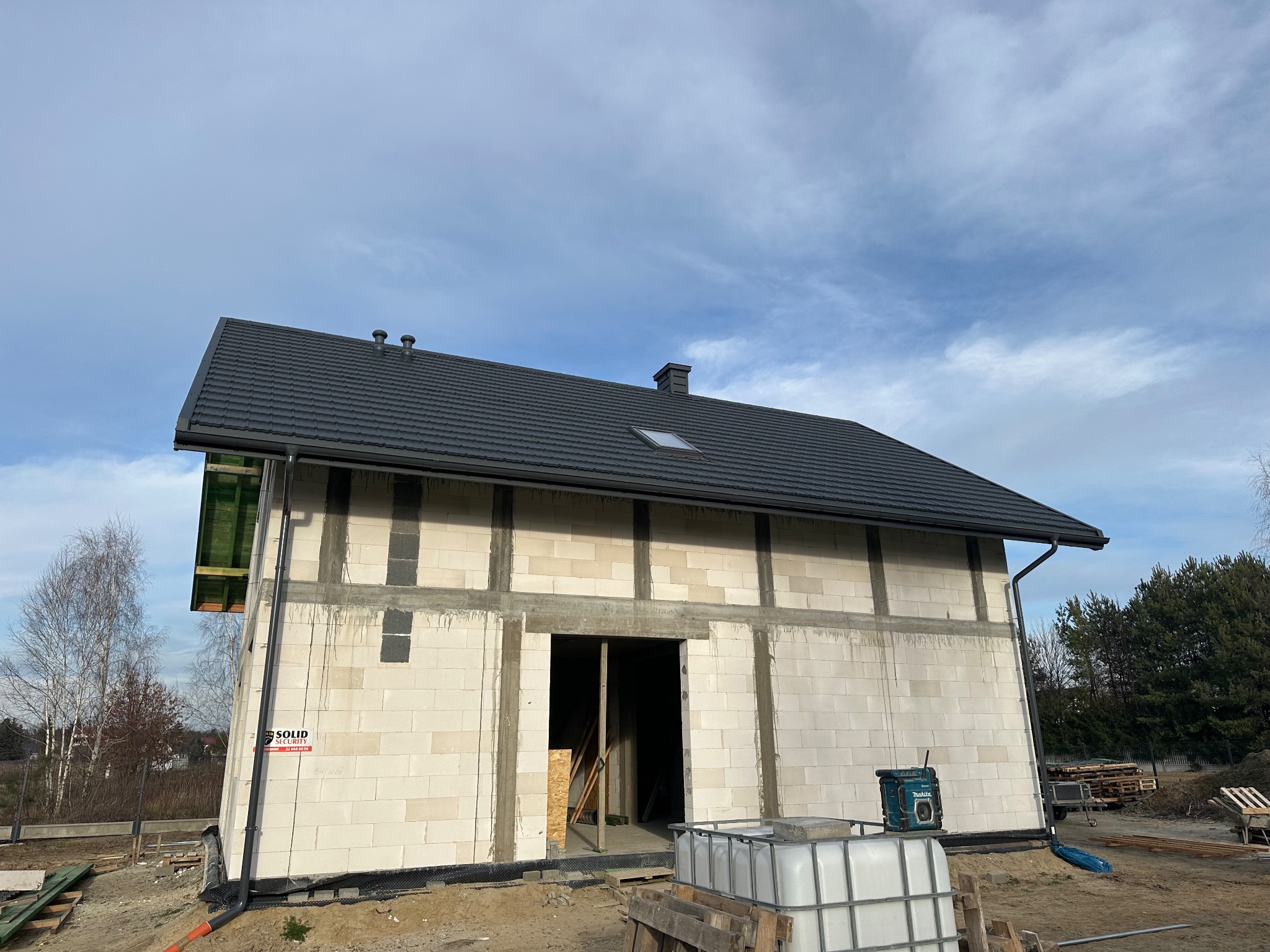 Surowy stan domu jednorodzinnego w trakcie budowy: widoczna czarna dachówka na skośnym dachu z oknem dachowym, zamontowane rynny, ściany z betonu komórkowego i niezabudowany otwór drzwiowy.