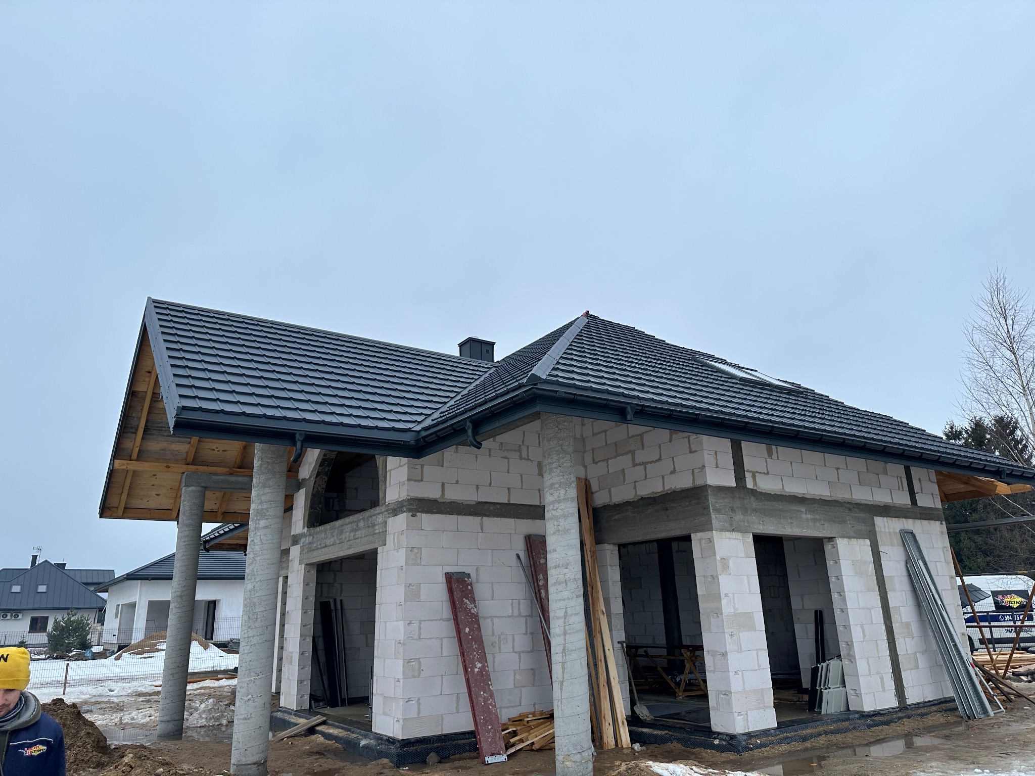 Dom w trakcie budowy z ciemną dachówką karpiówką i zamontowanymi rynnami. Widoczne bloczki betonowe, drewniane elementy konstrukcyjne i fragment osoby w żółtej czapce.