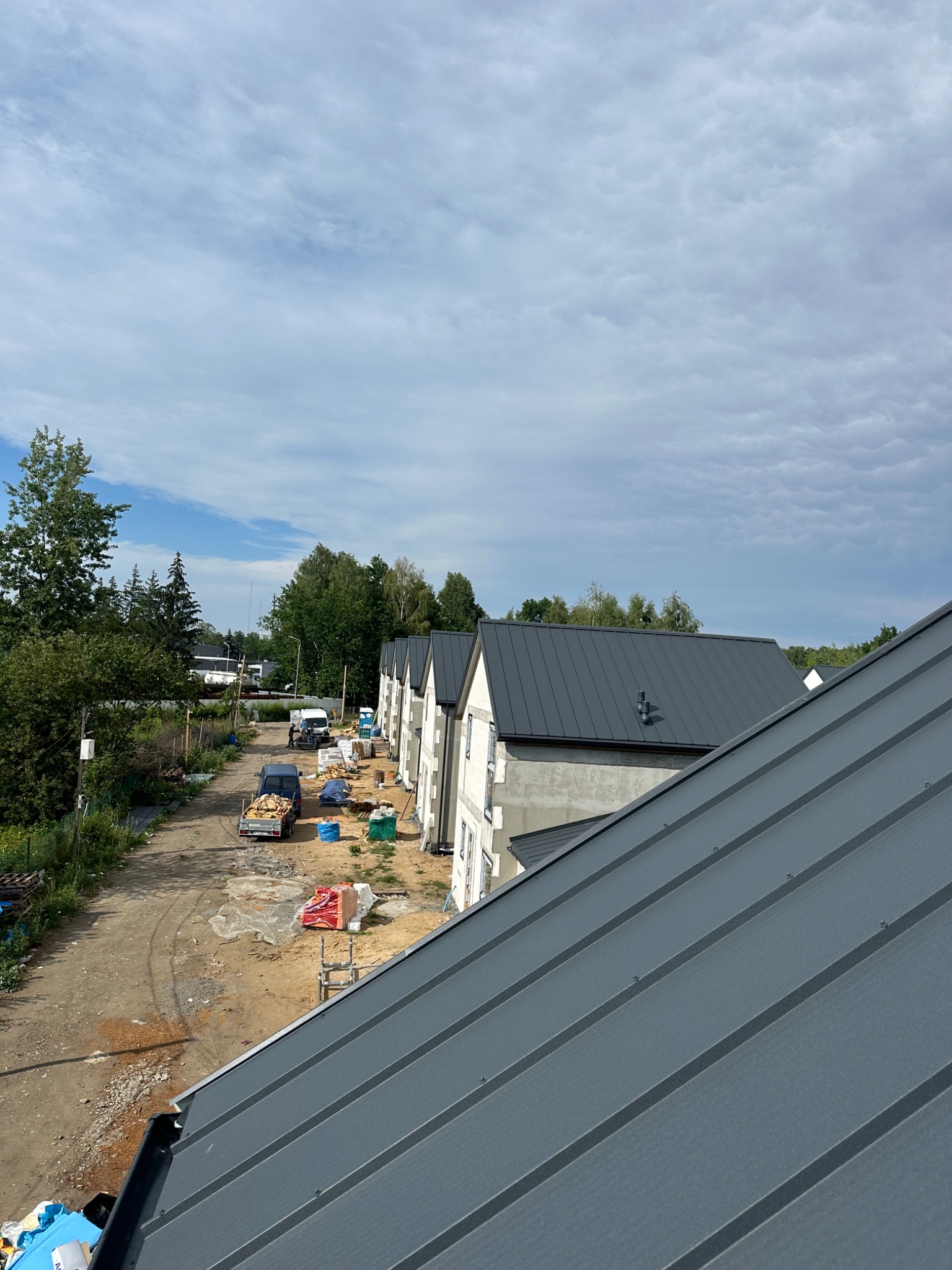 Szary dach z blachy na tle budynków w trakcie budowy, widok z góry. Na ziemi materiały budowlane i pojazdy. Pochmurne niebo.
