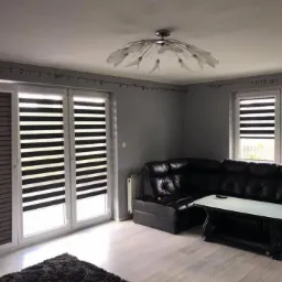 Salon z szarymi ścianami i dwiema roletami dzień noc w oknach, oraz jedną roletą wewnętrzną na przesuwanych drzwiach balkonowych. Na podłodze dywanik, a w rogu czarna skórzana kanapa.