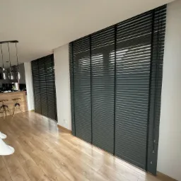 Wnętrze salonu z drewnianą podłogą i trzema oknami osłoniętymi ciemnymi żaluzjami poziomymi z szerokimi lamelami, widok na kuchnię z barkiem i hokerami.