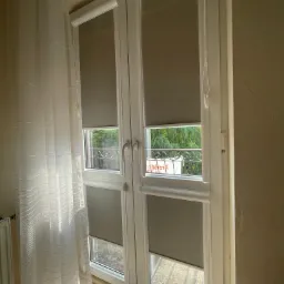 Dwu-skrzydłowe okno z szarymi roletami wewnętrznymi częściowo opuszczonymi, widok na balkon i zieleń za nim, po lewej stronie zwiewna biała firanka.