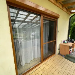 Zewnętrzna rama okienna z moskitierą, brązowe wykończenie, widok z zewnątrz na taras z kostki brukowej, obok stolik z narzędziami i akcesoriami.