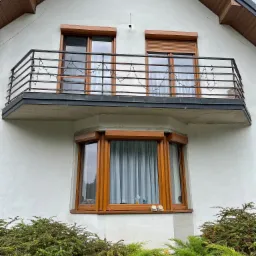 Fragment fasady domu z balkonem, na którym zamontowano brązowe rolety zewnętrzne, okna z brązowymi ramami, na parapecie dolnego okna stoją białe kapcie.