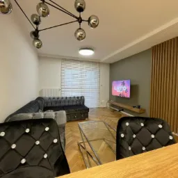 Nowoczesny salon z szarym narożnikiem Chesterfield, szklanym stolikiem kawowym i białymi żaluzjami poziomymi na oknie.