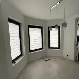 Jasne wnętrze z trzema oknami wyposażonymi w białe plisy poziome, wisząca lampa z trzema kloszami, szare płytki podłogowe, przewody na podłodze.