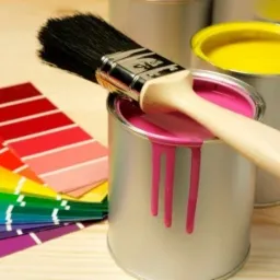 Pędzel zanurzony w puszce z różową farbą, obok paleta kolorów i puszka z żółtą farbą, na drewnianym podłożu.