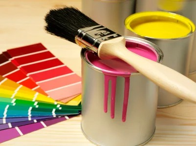 Pędzel zanurzony w puszce z różową farbą, obok paleta kolorów i puszka z żółtą farbą, na drewnianym podłożu.