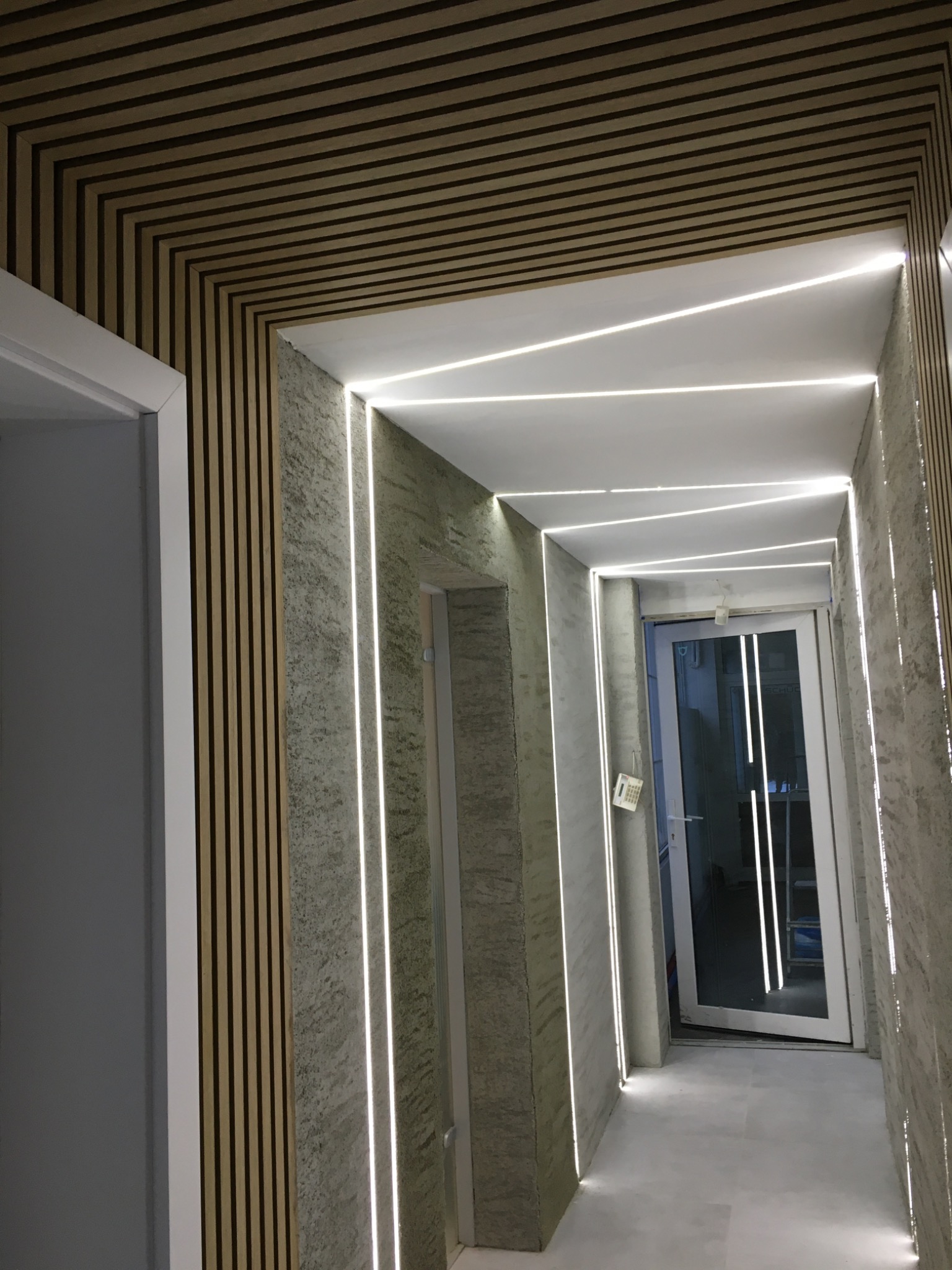 Korytarz z drewnianą boazerią na suficie i ścianach, oświetlony liniowymi lampami LED wzdłuż krawędzi ścian i sufitu, z widocznymi drzwiami na końcu korytarza.