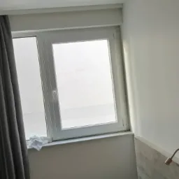 Białe okno PCV z szarymi zasłonami w jasnym pokoju, na parapecie leży zmięta chusteczka.