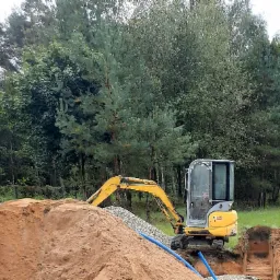 Żółta mini koparka E18 na tle lasu, obok wykopu z drenażem obsypanym żwirem i ułożonymi niebieskimi rurami.