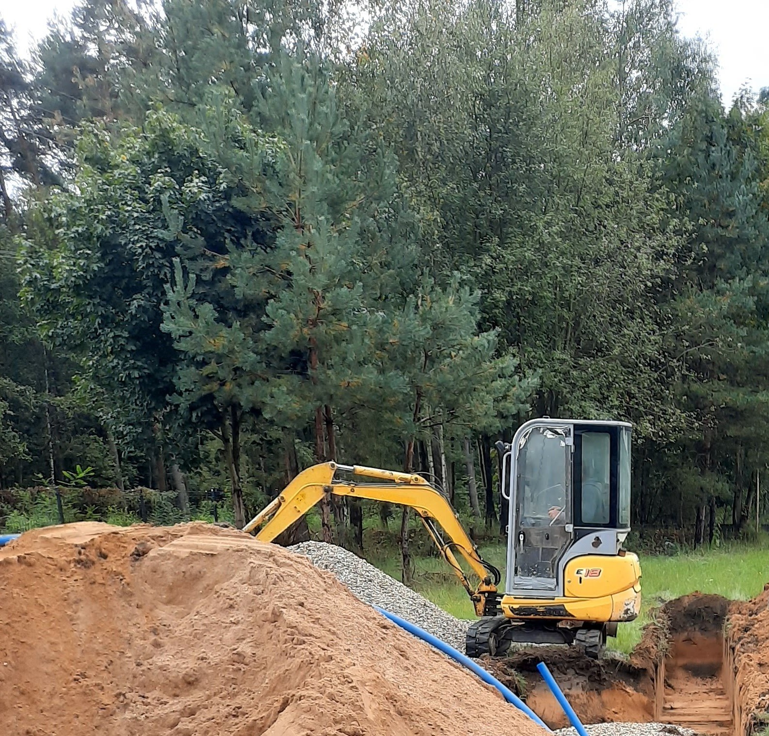 Żółta mini koparka E18 na tle lasu, obok wykopu z drenażem obsypanym żwirem i ułożonymi niebieskimi rurami.