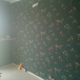 Poziom - Ściana w pokoju po tapetowaniu, widoczny wzór w ptaki i kwiaty na ciemnozielonym tle, fragment sufitu z listwą dekoracyjną, gniazdko elektryczne z wystającymi kablami, żółta szpachelka oparta...