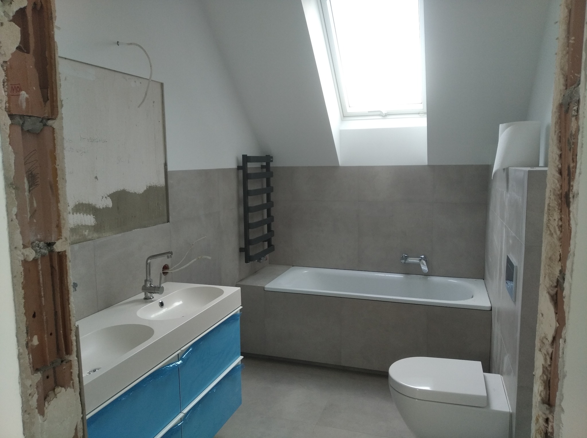 Łazienka na poddaszu w trakcie remontu: widoczna wanna w zabudowie z szarych płytek, umywalka z szafką zabezpieczoną niebieską folią, grzejnik drabinkowy i toaleta, ściany częściowo wykończone...