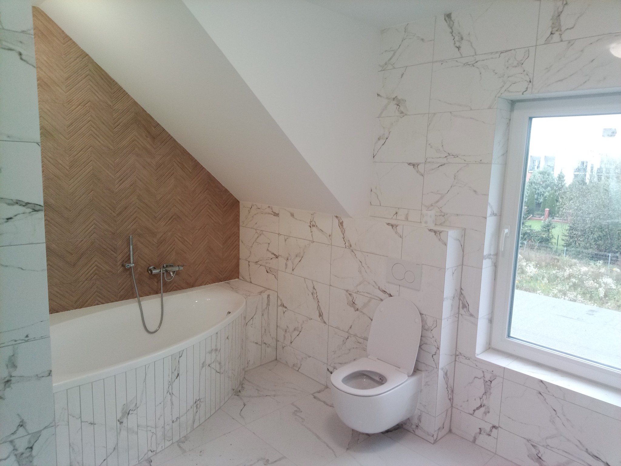 Nowoczesna łazienka na poddaszu z wanną obudowaną marmurowymi płytkami, toaletą podwieszaną i ścianą z drewnopodobną okładziną w jodełkę.